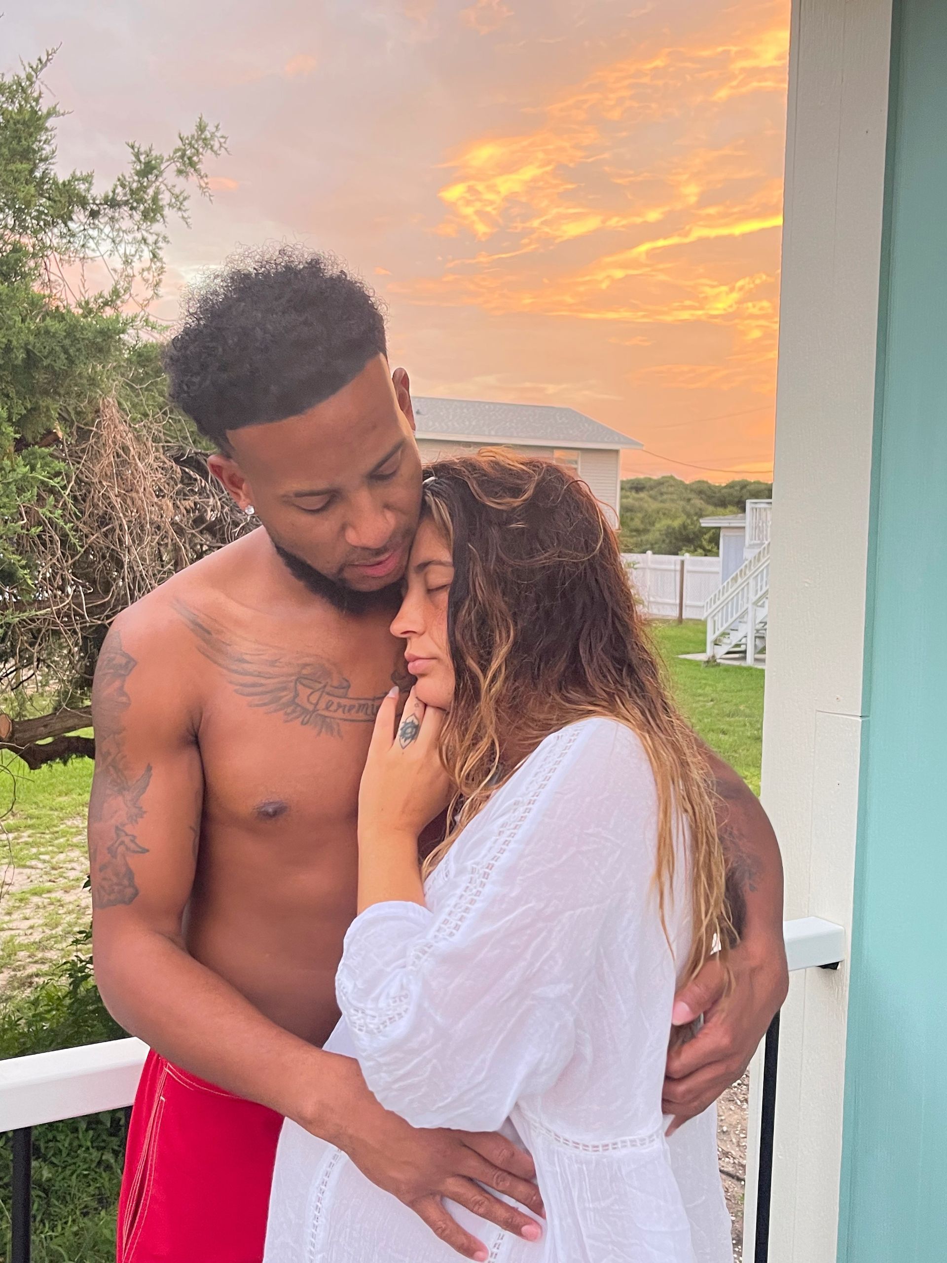 Couple embracing on a porch at sunset, with warm orange sky and greenery in the background.