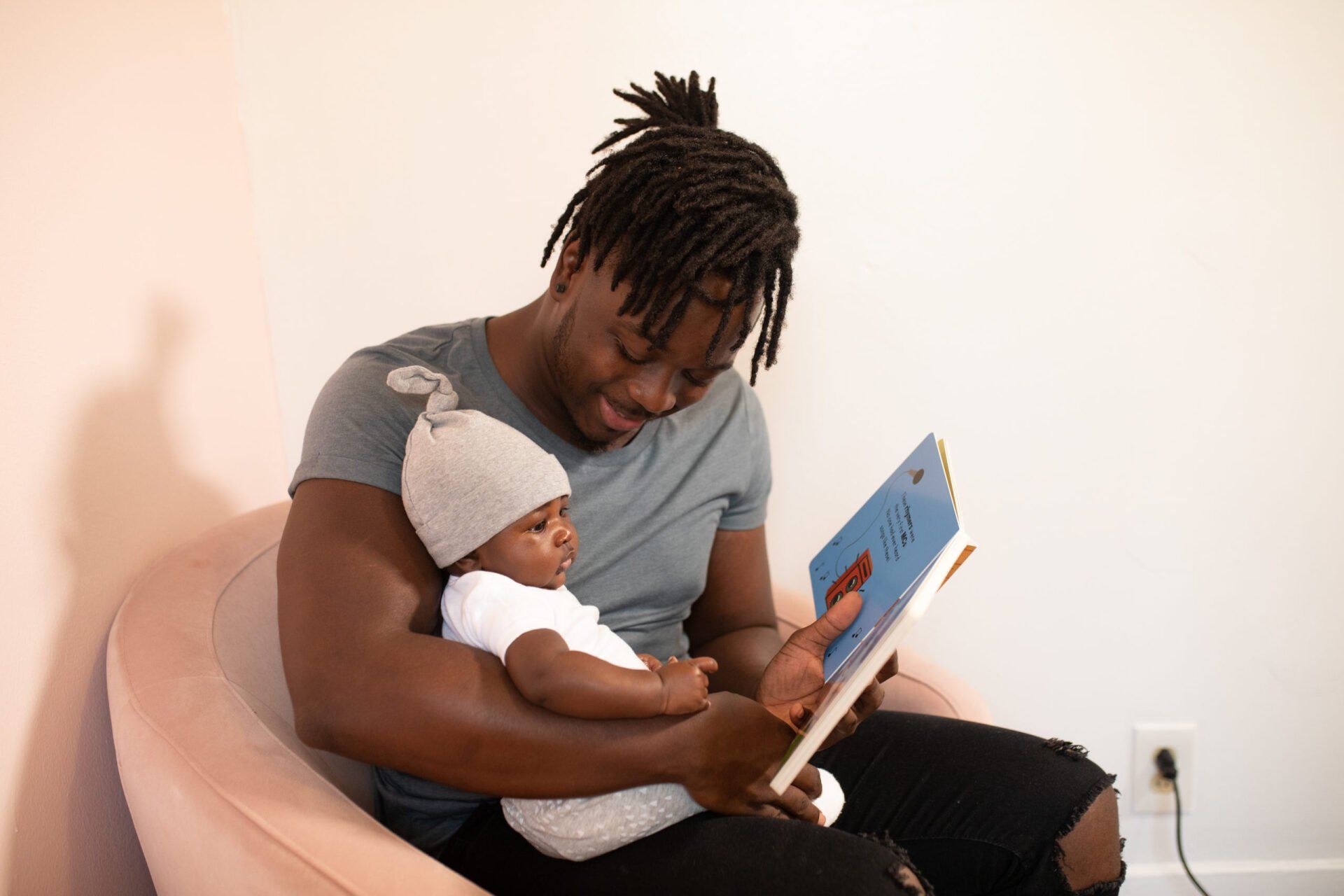 Adult reading a book to a baby on a pink chair in a bright room