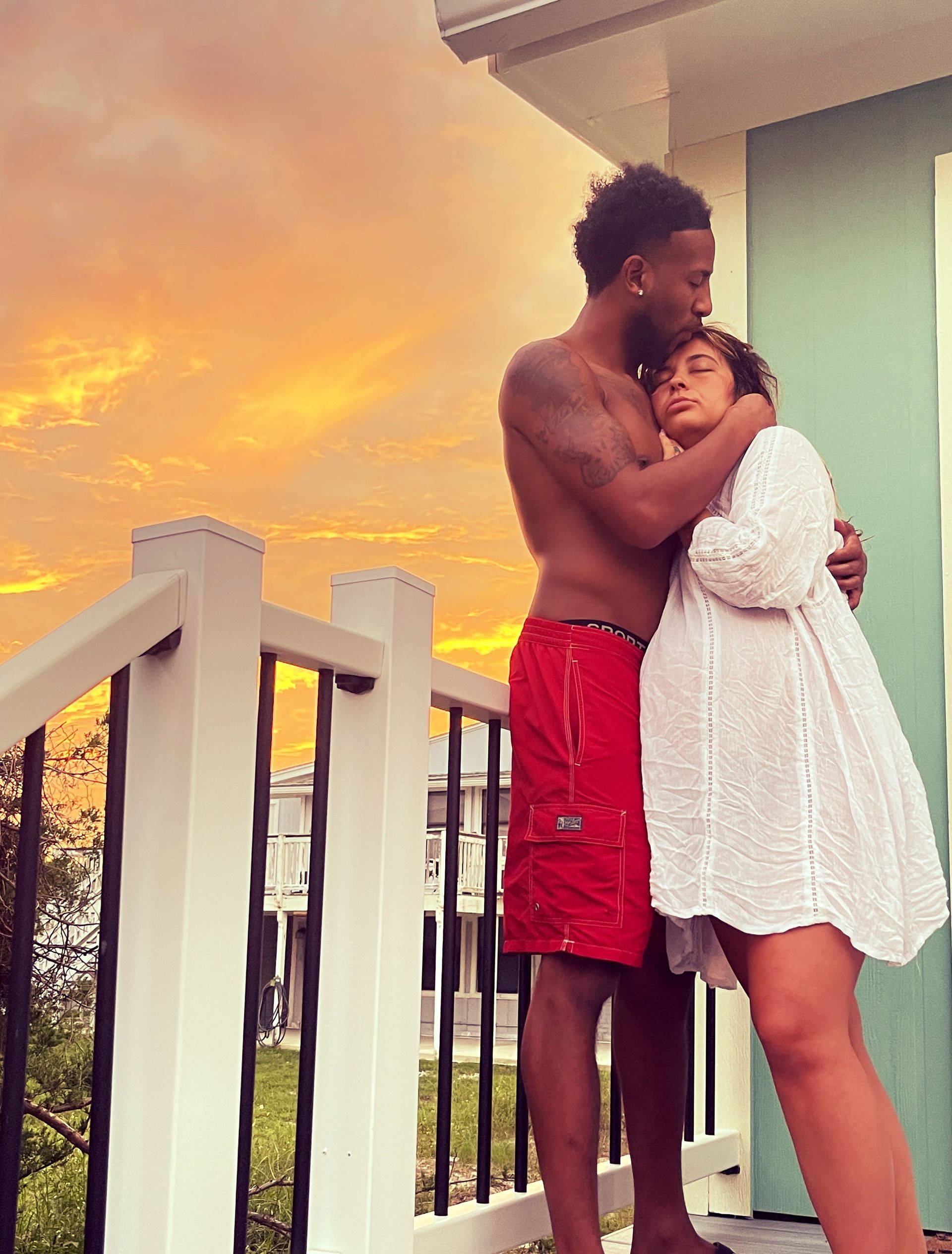 Couple embracing on a balcony at sunset, with orange sky and white railings.
